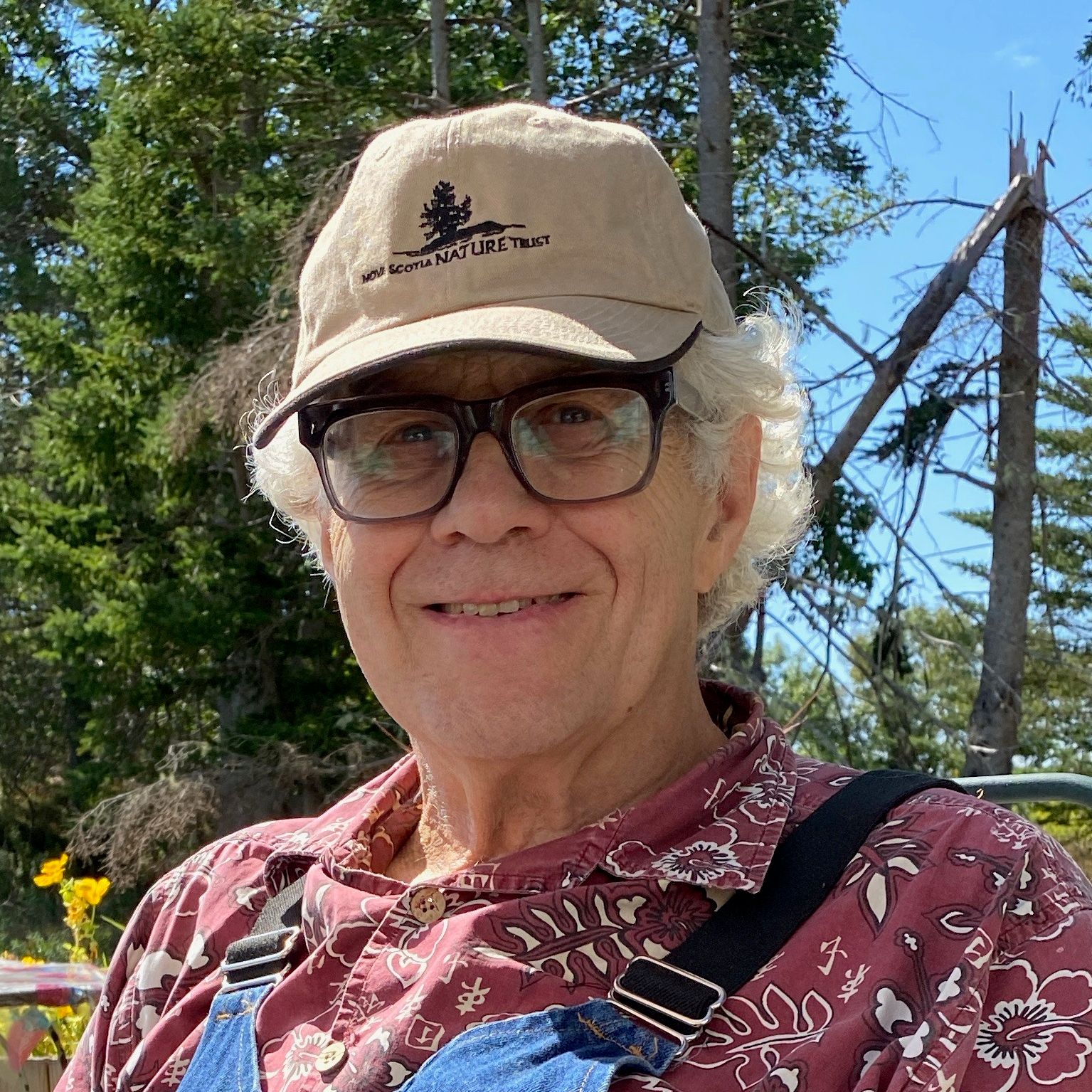 A man with thick-rimmed glasses and curly white hair, wearing a tan ballcap, a red and white Aloha shirt and blue denim overalls, smiles in front of spruce trees and yellow flowers.